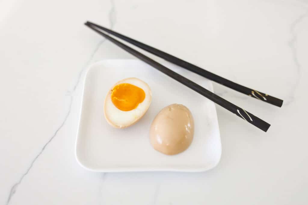 split in half ajitama egg show on a plate with chopsticks. One half is face up to show the perfect yolk and the other is face down to show the coloration.