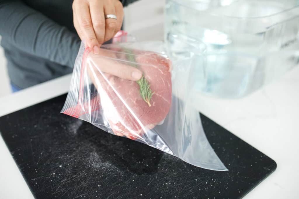 Sirloin steak and rosemary sprig being placed in a Ziploc bag.