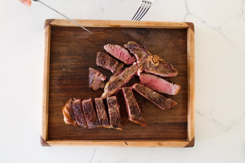 Overhead shot of a nicely charred porterhouse carved on a wooden tray ready to eat. A piece of tenderloin and strip steak is flipped sideways so you can see it's perfectly cooked.