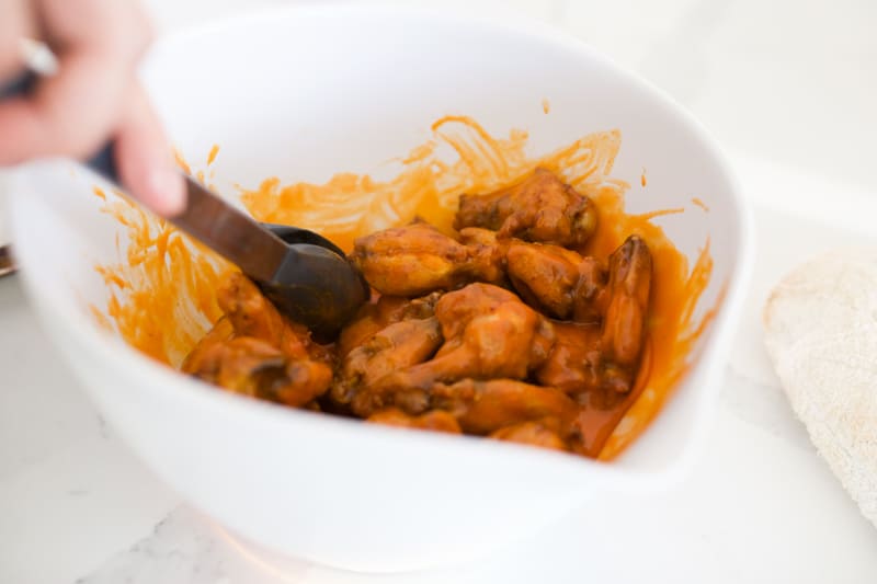 Chicken wings being tossed with tongs in buffalo sauce.