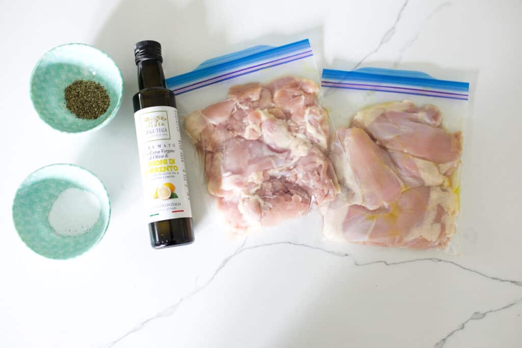 Salt and pepper in bowls, a bottle of lemon infused olive oil, and two bags of chicken thighs in Ziploc freezer bags.