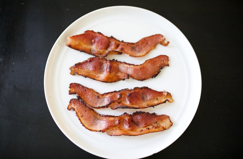 Four strips of bacon on a white plate on a black table.